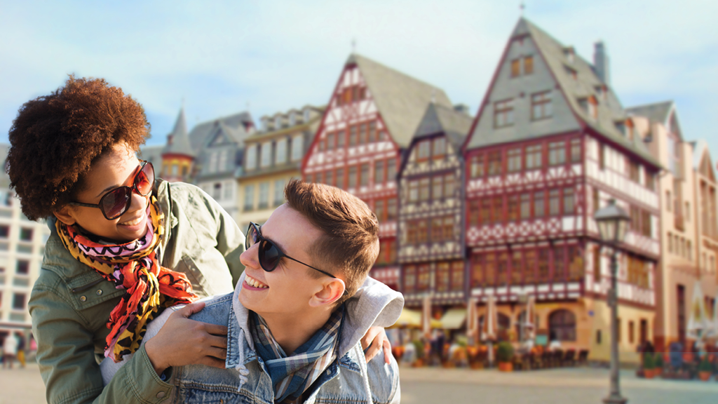 A photo of a man and woman wearing sunglasses smiling at each other. Behind them is a traditional-looking building in Frankfurt.