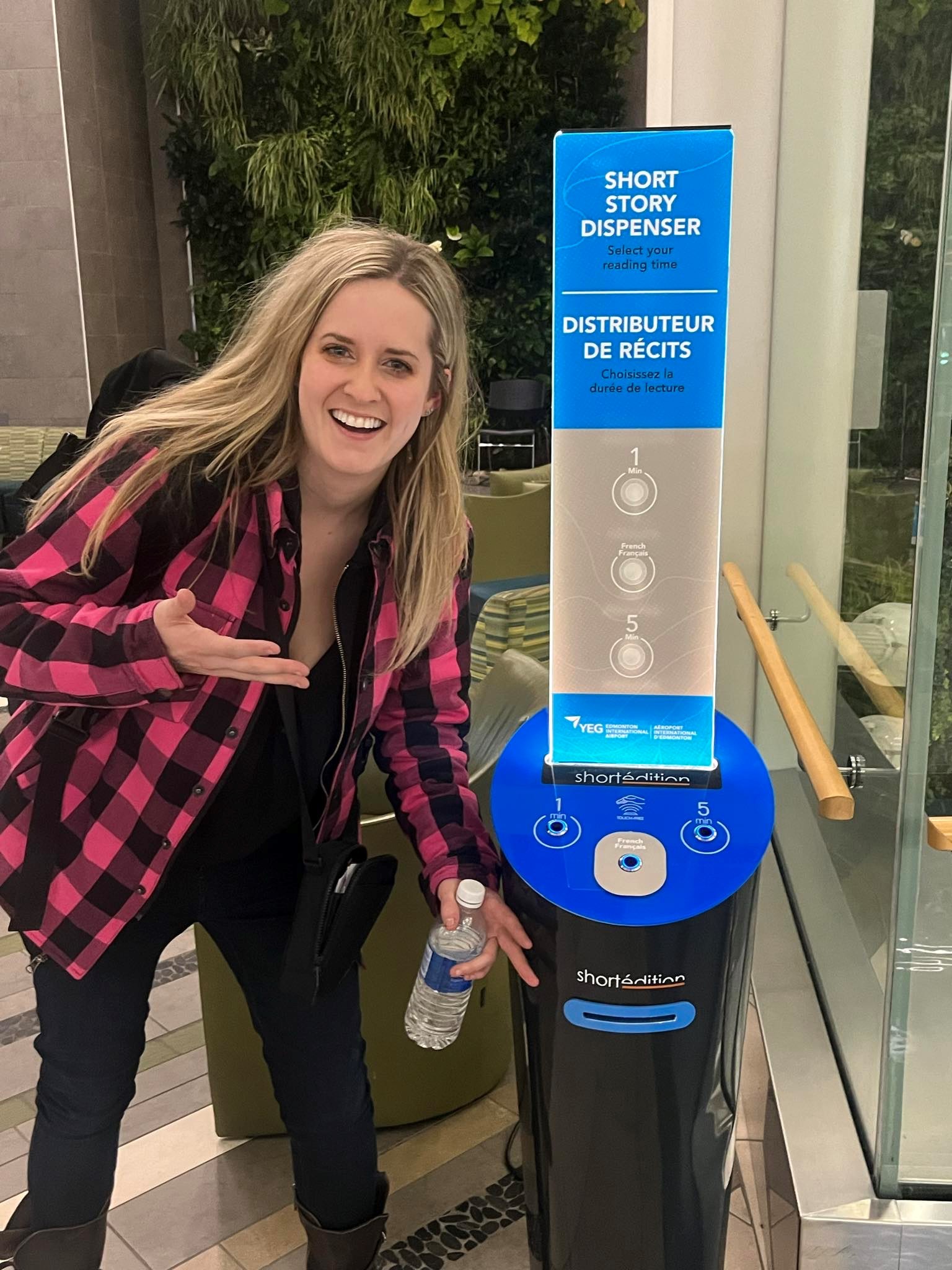 Smiling woman in plaid shirt points to the Short Story Dispenser at Edmonton International Airport